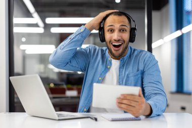 Masada oturan, kulaklık takan, tablet kullanan, elini kafasına dayayıp kameraya gülümseyen mutlu genç Müslüman bir adamın portresi..