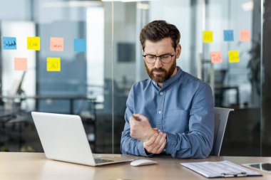 Yorgun genç bir adam ofiste fazla mesai yapıyor, elinde dizüstü bilgisayarla masada oturuyor, elini bileğinden tutuyor, masaj yapıyor, şiddetli bir acı hissediyor..