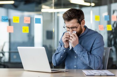 Virüs ve grip hastası genç bir adam modern bir ofiste bir masada oturuyor ve burnundan akan bir peçeteyle burnunu siliyor..