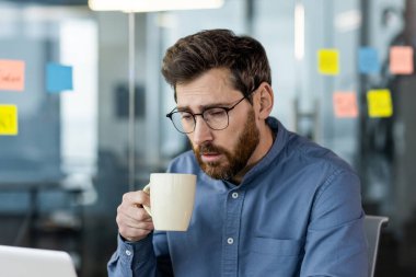 Ofiste kahve fincanı tutan, gözlük takan, dizüstü bilgisayara bakan yorgun işadamı. Modern iş ortamında stres, yorgunluk ve çalışma baskısı kavramı