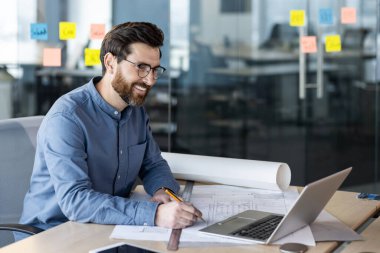 Genç, gülümseyen bir mimar, mühendis ve müteahhit çizimlere göre ofiste çalışıyor, elinde belgelerle bir masada oturuyor ve bir video görüşmesi yapıyor..