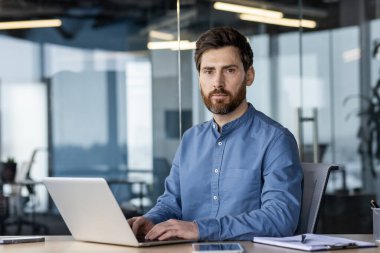 Ofiste çalışan genç bir adamın portresi dizüstü bilgisayarın başında oturuyor ve kendinden emin bir şekilde kameraya bakıyor..