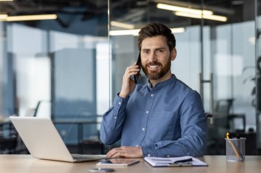 Modern bir ofiste çalışan gülümseyen genç bir adamın portresi, bir masada dizüstü bilgisayarla oturuyor, telefonla konuşuyor ve kameraya bakıyor..
