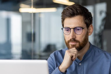Gözlüklü düşünceli bir gencin yakın plan fotoğrafı. Laptop ekranına bakıyor, sakalına elini uzatıyor ve ciddi ciddi bir çözüm düşünüyor..