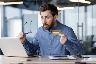 Şok olmuş genç bir ofis çalışanı iş yerinde, elinde cüzdan ve kredi kartıyla dizüstü bilgisayar ekranına endişeli bir şekilde bakıyor..