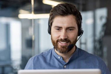 Mavi gömlekli genç bir adamın, iş yerinde kulaklıkla otururken ve dizüstü bilgisayarla çalışırken çekilmiş yakın plan fotoğrafı..