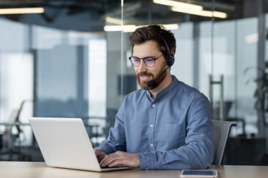 Mavi gömlekli gülümseyen genç bir adam modern bir ofiste bir masada oturuyor, kulaklık takıyor, bilgisayarında çalışıyor ve çalışıyor..