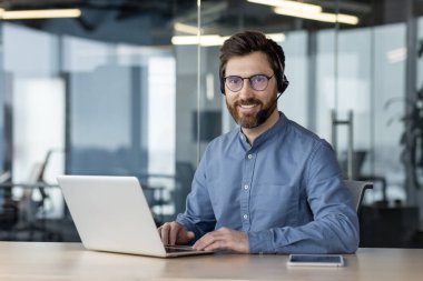 Ofiste dizüstü bilgisayarla oturmuş kameraya güvenle bakan, kulaklık takan gülümseyen bir gencin portresi..