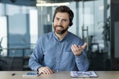 Ofisteki masada oturmuş kameraya elini uzatan mavi gömlekli gülümseyen genç bir adamın portresi..