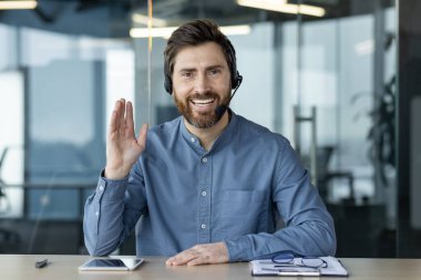 Kulaklık takmış gülümseyen genç bir adam ofiste oturur, konuşur, gülümser ve elini kameraya uzatır, çevrimiçi danışmanlık yapar ve uzaktan eğitim verir..