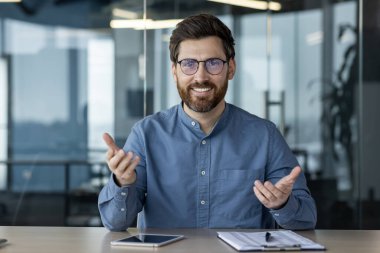 İnternetteki konferans ve iş toplantısı. Gözlüklü, gülümseyen genç bir adam kameranın önünde oturur ve bir video görüşmesinde elleriyle işaret eder..