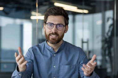 Gözlüklü ve mavi gömlekli genç bir adamın yakın plan portresi. Ofiste kameranın önünde oturuyor ve bir video konuşmasında elleriyle işaret ediyor..