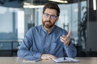 Mesafe öğrenme. Ofisteki masada oturan genç ve ciddi bir adam online bir konferans, iş eğitimi ve toplantı yürütüyor. Kameraya bak ve konuş..