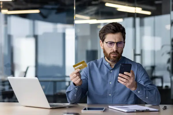 stock image Upset young male businessman worried about credit card and mobile phone problem, sitting in office at desk.