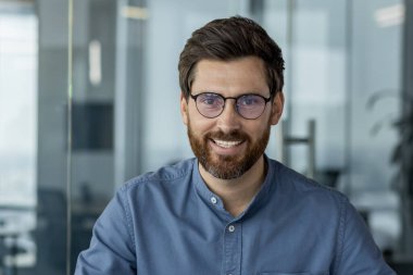 Gözlüklü ve mavi gömlekli genç bir adamın yakın plan portresi ofiste oturuyor ve kameraya özgüvenle bakıyor..