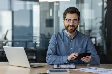 Modern ofis ortamında tablet ve laptop kullanan gülümseyen bir işadamı. Profesyonel çalışma ortamı, cam duvarlar ve masa başında evrak işleri. İş, teknoloji ve işyeri kavramı