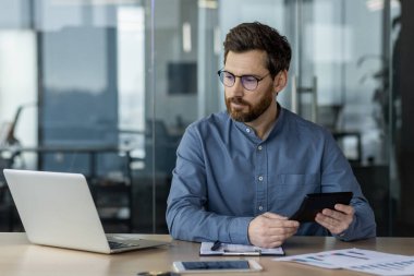 Modern ofis ortamında dizüstü bilgisayar ve tablet kullanan ciddi bir muhasebeci. Evrak işleri, veriler ve hesaplamalara odaklanmış. Üretkenlik, finans ve profesyonelliği kavramsal