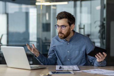Hüsrana uğramış işadamı dizüstü bilgisayar ve tabletle masada oturuyor, kafa karışıklığı ve stres gösteriyor. Modern ofis ortamında işyeri sorunları, çoklu görev ve sorun çözme kavramı