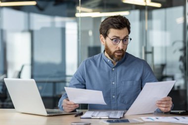 Gözlüklü, şok olmuş genç bir adam elinde dizüstü bilgisayarla ofiste oturuyor ve elindeki belgelere ve belgelere endişeli bir şekilde bakıyor..