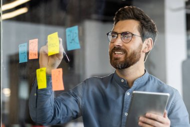 İş adamı, elinde tablet tutarken yapışkan notlar kullanarak cam panoda beyin fırtınası yapıyor ve fikirler planlıyor. Modern ofis ortamında yaratıcı süreçlere ve takım çalışmalarına bağlıdır.