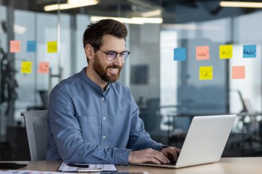 Gülümseyen genç bir işadamı masadaki ofiste oturuyor ve bir dizüstü bilgisayar üzerinde çalışıyor..