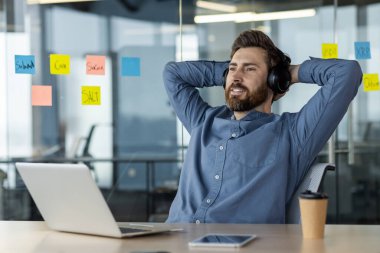 Gülümseyen ve huysuz kulaklıklı genç bir adam iş yerinde dizüstü bilgisayarın önünde bir sandalyede oturuyor ve elleri başının arkasında dinleniyor..