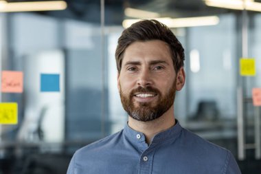 Modern ofiste gülümseyen sakallı, kendinden emin bir iş adamı. Profesyonel, mavi gömlekli, iş ortamında takılıyor. Ofis cam duvarları ve arka planda renkli yapışkan notlar.