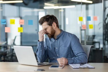 Mavi gömlekli yorgun genç bir adam ofisteki masada oturuyor ve laptopta fazla mesai yapıyor, elleriyle gözlerini ovuyor, gözlük tutuyor..