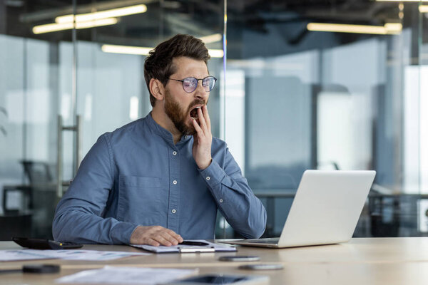 Уставший молодой человек, офисный работник сидит в офисе сверхурочно, работает на ноутбуке, зевает, закрывает рот рукой.