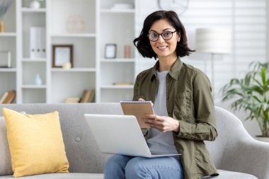 Gözlüklü ve günlük kıyafetlerle gülümseyen genç bir kadının portresi. Evdeki kanepede dizüstü bilgisayarın önünde oturuyor. Not defterine not alıyor ve kameraya bakıyor..