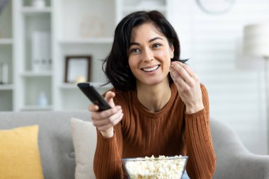 Gülümseyen genç bir kadın TV karşısında dinleniyor, koltukta oturuyor, patlamış mısır yiyor ve uzaktan kumanda kullanıyor. Yakın plan fotoğraf..