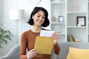 Sıcak bir oturma odasında mektup okuyan neşeli bir kadın. Mutlu bir haber ya da davetiye alma anı.