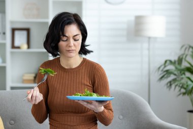 Modern bir oturma odasında bir tabak salata tutan düşünceli bir kadın yemek seçimini düşünüyor..