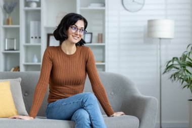 Genç bir kadın evdeki kanepede oturuyor, gözlük ve kahverengi bir kazak giyiyor, yan tarafa bakıyor ve gülümsüyor..