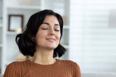 Evdeki genç ve güzel esmer kadının yakın plan fotoğrafı. Kanepede gözleri kapalı, dinleniyor, gülümsüyor ve derin nefes alıyor..