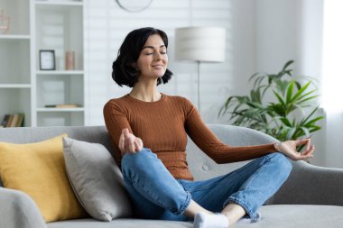 Nilüferli genç kız yoga yapıyor, evdeki kanepede oturuyor, gözleri kapalı meditasyon yapıyor..