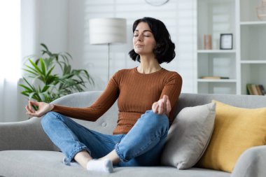 Güzel, genç, rahat bir kadın evde yoga yapıyor, gözleri kapalı bir şekilde Lotus pozisyonunda oturuyor, meditasyon yapıyor ve dinleniyor..