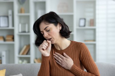 Evdeki genç bir kadının yakın plan fotoğrafı, kanepede oturuyor ve öksürüyor, göğsünde ağrı hissediyor, eliyle ağzını kapatıyor..