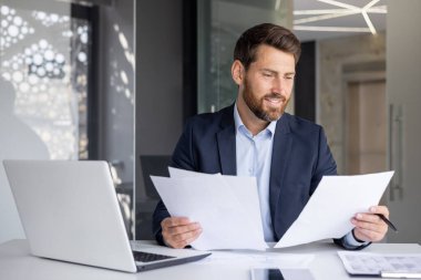 Ofiste belgelerle çalışan gülümseyen genç bir adam elinde dizüstü bilgisayarla bir masaya oturur ve elindeki kağıtlara bakar..