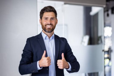 Koyu takım elbiseli profesyonel ve kendine güvenen bir yönetici iyi ışıklandırılmış, modern ofis ortamında iyi bir jest yapıyor, pozitiflik ve liderlik yayıyor..