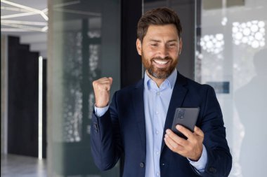 Modern ofis ortamında neşeli bir işadamı akıllı telefonuna bakarken başarıyı kutluyor. Takım elbise giyiyor ve kendine güveni tam, mutlu bir ifadesi var..