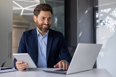 Şık takım elbiseli kendine güvenen bir işadamı modern ofis ortamında dizüstü bilgisayar ve tablet kullanarak başarılı bir şekilde çalışır..