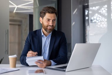 Modern bir ofiste profesyonel orta yaşlı bir iş adamı. İşe dalmış, dizüstü bilgisayar kullanıyor ve gülümseyen bir tavırla notlar yazıyor..