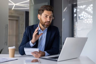 Modern bir ofiste profesyonel bir erkek işadamı görüntülü görüşme yaptı. Laptop kullanırken ve notları tutarken aktif olarak tartışıyor ve hareketler yapıyor..