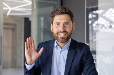 30 'lu yaşlarda, takım elbiseli profesyonel bir erkek modern ofis ortamında neşeli bir gülümsemeyle selamlaşır..