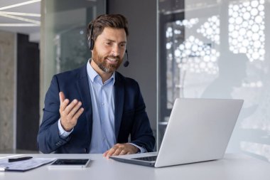 Mavi takım elbiseli ve kulaklıklı profesyonel bir erkek modern bir ofiste online bir iş toplantısında laptopun başında tutkuyla konuşuyor..