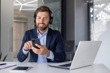 Neşeli bir işadamı akıllı telefonuyla modern ofis ortamında kulaklık kullanırken rahat ve profesyonel görünmeye çalışır..