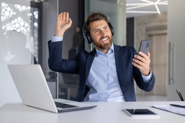 Takım elbiseli gülümseyen işadamı kulaklıkla müzik dinliyor, modern bir ofiste akıllı bir telefonu tutarken elini neşeyle kaldırıyor..