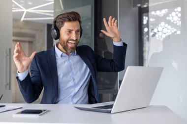 Takım elbiseli ve kulaklıklı neşeli bir işadamı, modern bir ofiste yapılan bir video görüşmesi sırasında anlamlı jestlerle kutlama yapıyor. İmaj başarıyı, heyecanı ve profesyonelliği yansıtıyor..