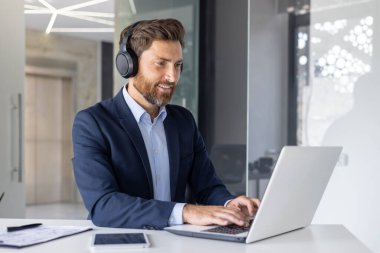 Kulaklık takan neşeli bir işadamı modern bir ofis masasında oturmuş bir dizüstü bilgisayarın üzerinde çalışıyor ve şirket ortamında odak ve profesyonelliği canlandırıyor..